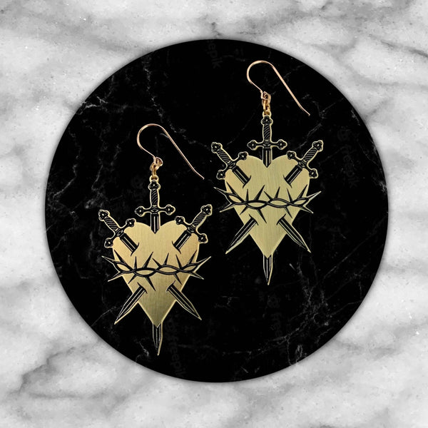 Gold heart-shaped earrings with sword designs on a black marble background
